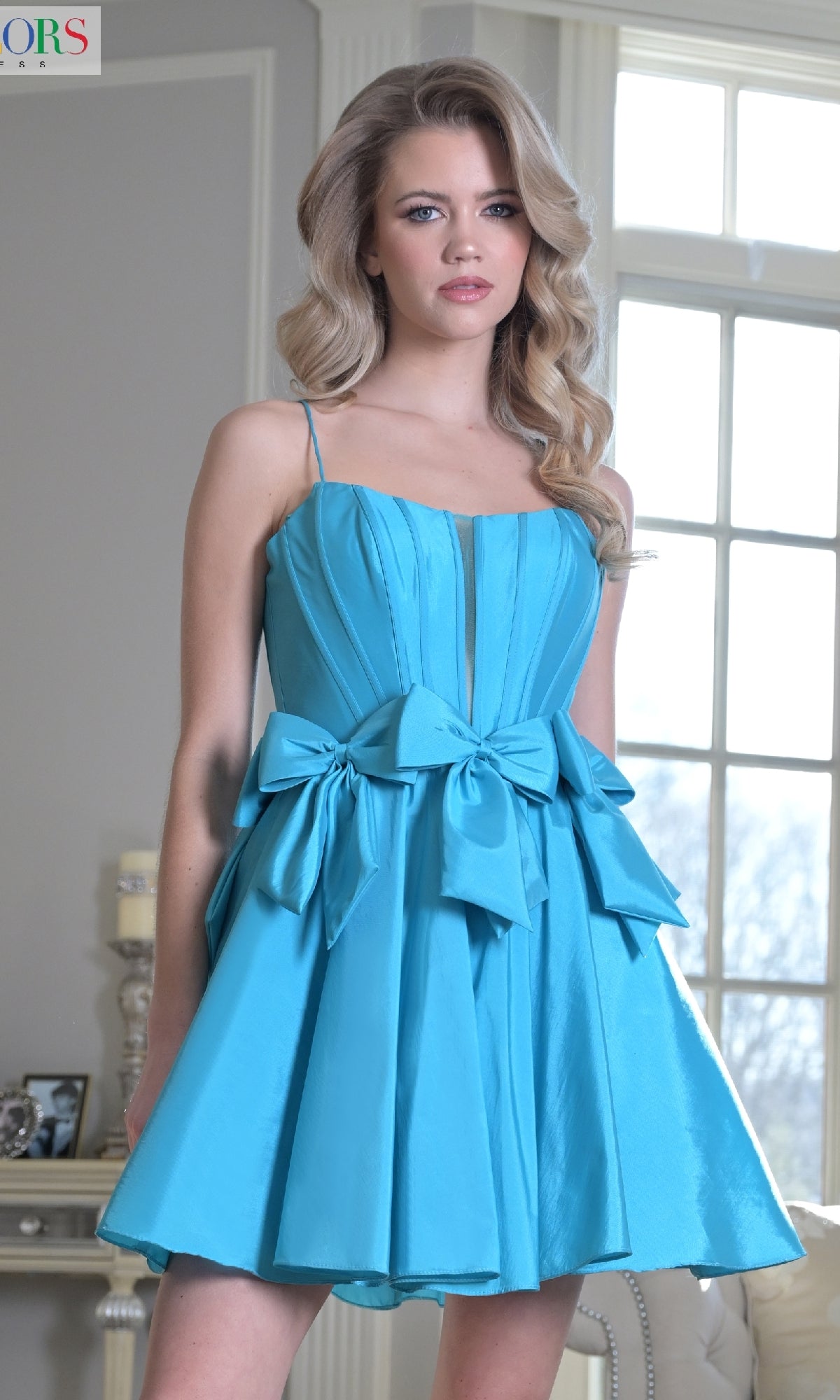 A woman wears the Bow-Waist Short Hoco Dress: Colors Dress 3637—a blue, sleeveless dress with a corset-style bodice and two large waist bows—standing indoors before large windows.